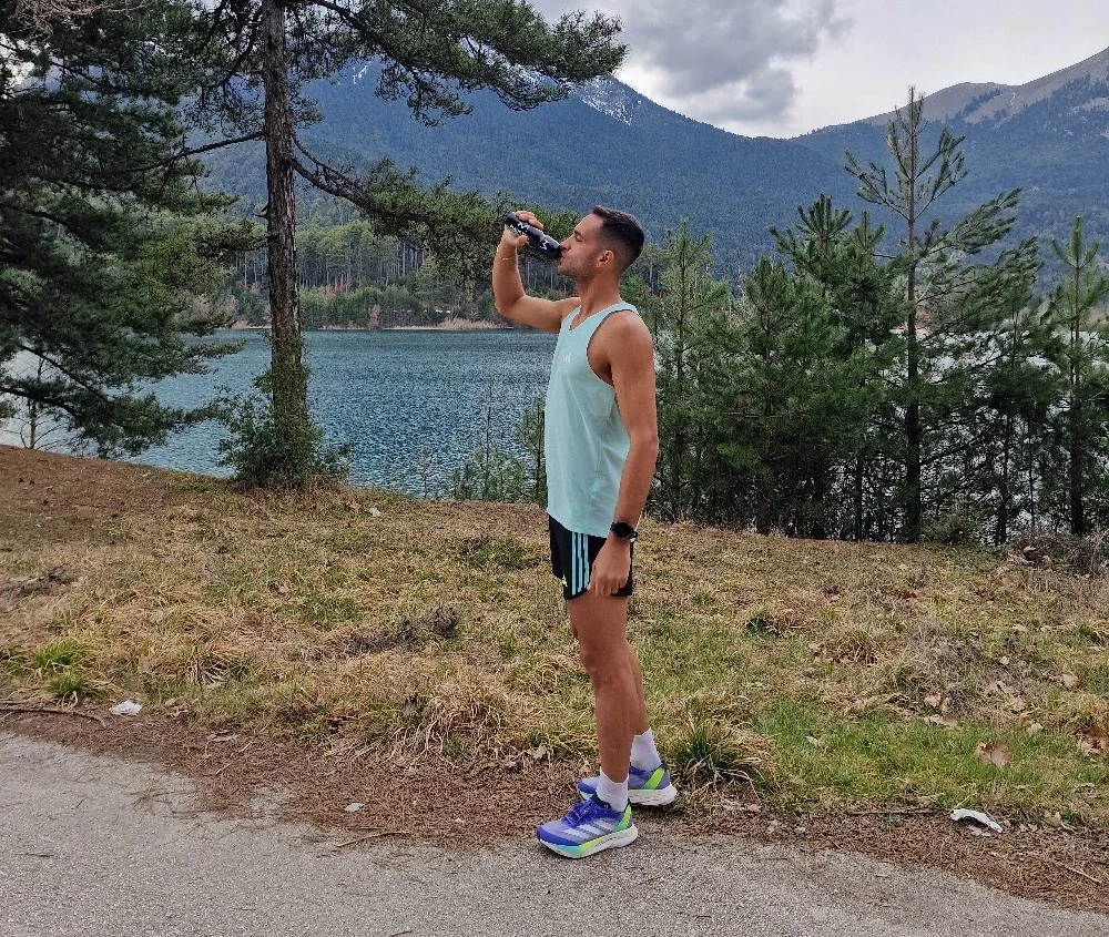 Δρομέας πίνει νερό δίπλα σε λίμνη με φόντο βουνά.